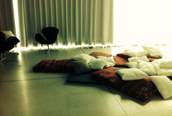Ein Raum, der durch Vorhänge abgedunkelt werden kann. Im Vordergrund eine schwarze dünne Matratze mit vielen roten und weißen Kissen. Im Hintergrund zwei gemütliche Drehstühle. Unter dem Fenster liegen Rosenblüten.