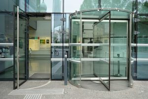 Blick auf den Eingang des Universalmuseums Joanneum in Graz, Österreich. Rechts befindet sich eine Drehtür aus Glas. links ein separater Eingang für Menschen mit Rollstuhl. Ein Knopf zum automatischen Türöffnen ist sichtbar. Auf dem Boden befinden sich taktile Blindenleitsysteme.
