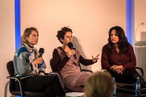 Ein Podium einer Veranstaltung der Berliner Gazette in Berlin. Auf dem Podium sitzen drei Frauen, zwei haben eine Migrationsgeschichte.