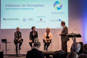 Ein Podium auf der Veranstaltung "Inklusion im Fernsehen" von der Grimme Akademie bei RTL in Köln. Auf dem Podium sitzen drei Frauen, zwei blonde Frauen und eine dunkelhaarige Frau. Eine der Frauen ist blind. Neben ihnen steht der Moderator.